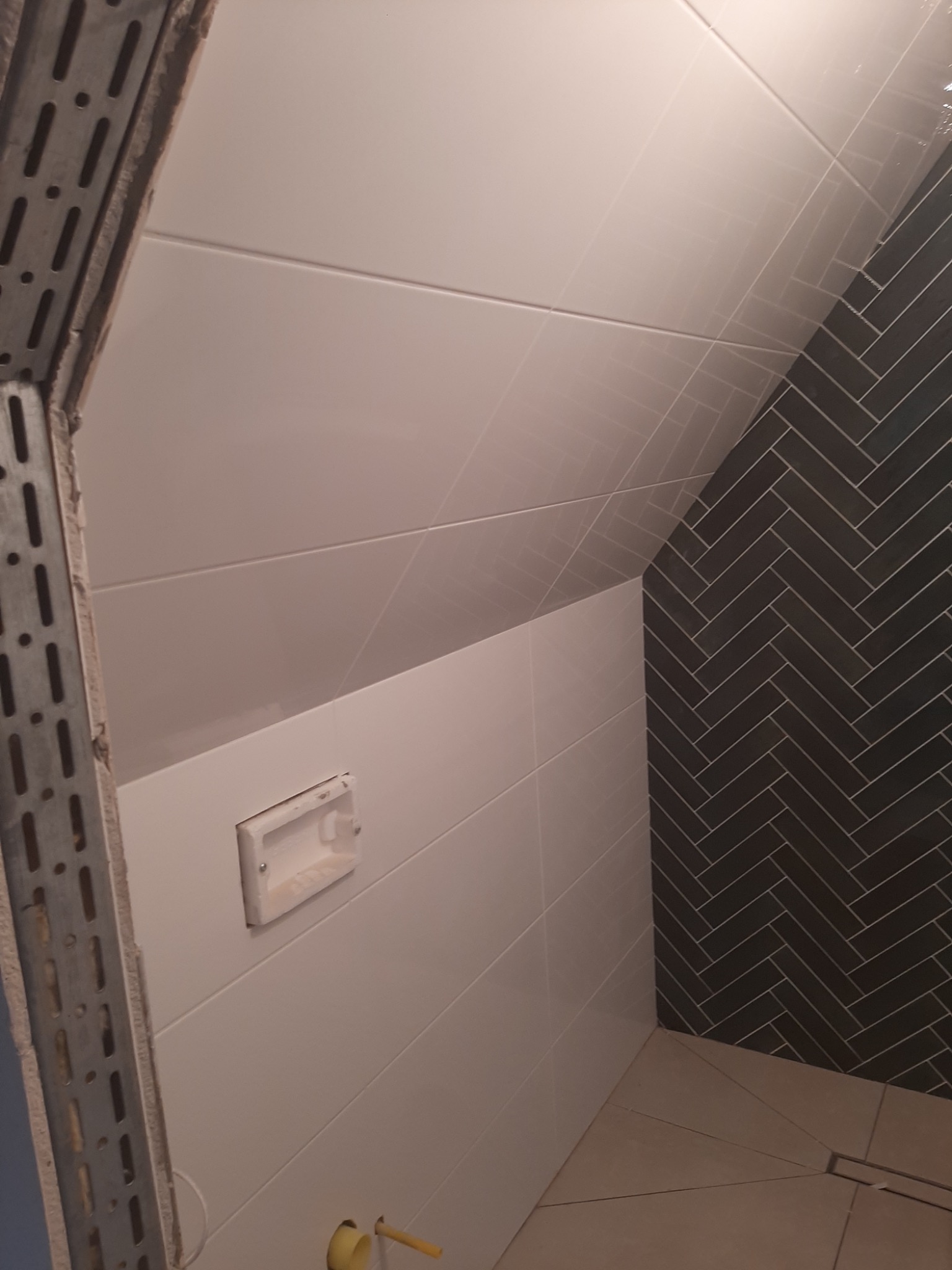 Ściana w łazience wyłożona białymi, poziomymi płytkami z widoczną wnęką na akcesoria oraz fragment ściany wyłożonej ciemnymi płytkami w jodełkę, widoczny metalowy profil przy krawędzi ściany.