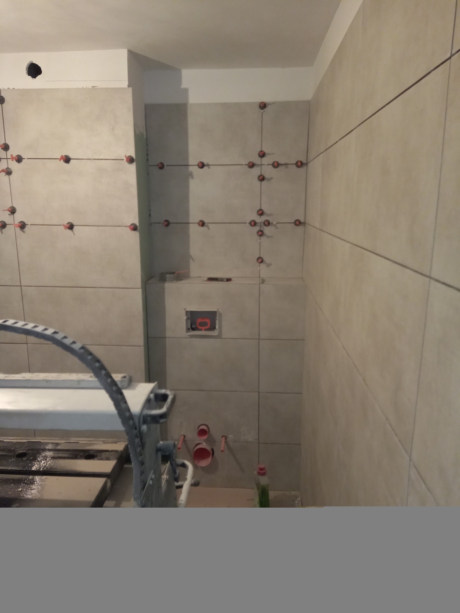 Ściana łazienki w trakcie układania płytek ceramicznych, widoczne krzyżyki dystansowe, poziomica laserowa, otwór na stelaż WC i obcięte rury kanalizacyjne.