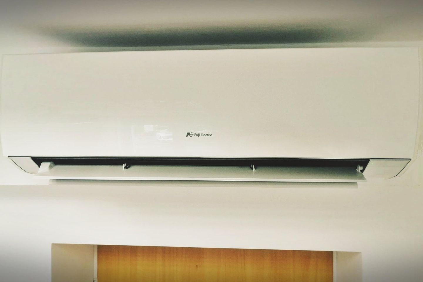 Biała jednostka wewnętrzna klimatyzacji Fuji Electric zamontowana na ścianie, z widocznym logo i otwartą klapką nawiewu.