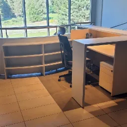 Jasne biuro z recepcją w kształcie litery L, regałem z półkami i szafkami zamykanymi na klucz, widok na zieleń za oknem.