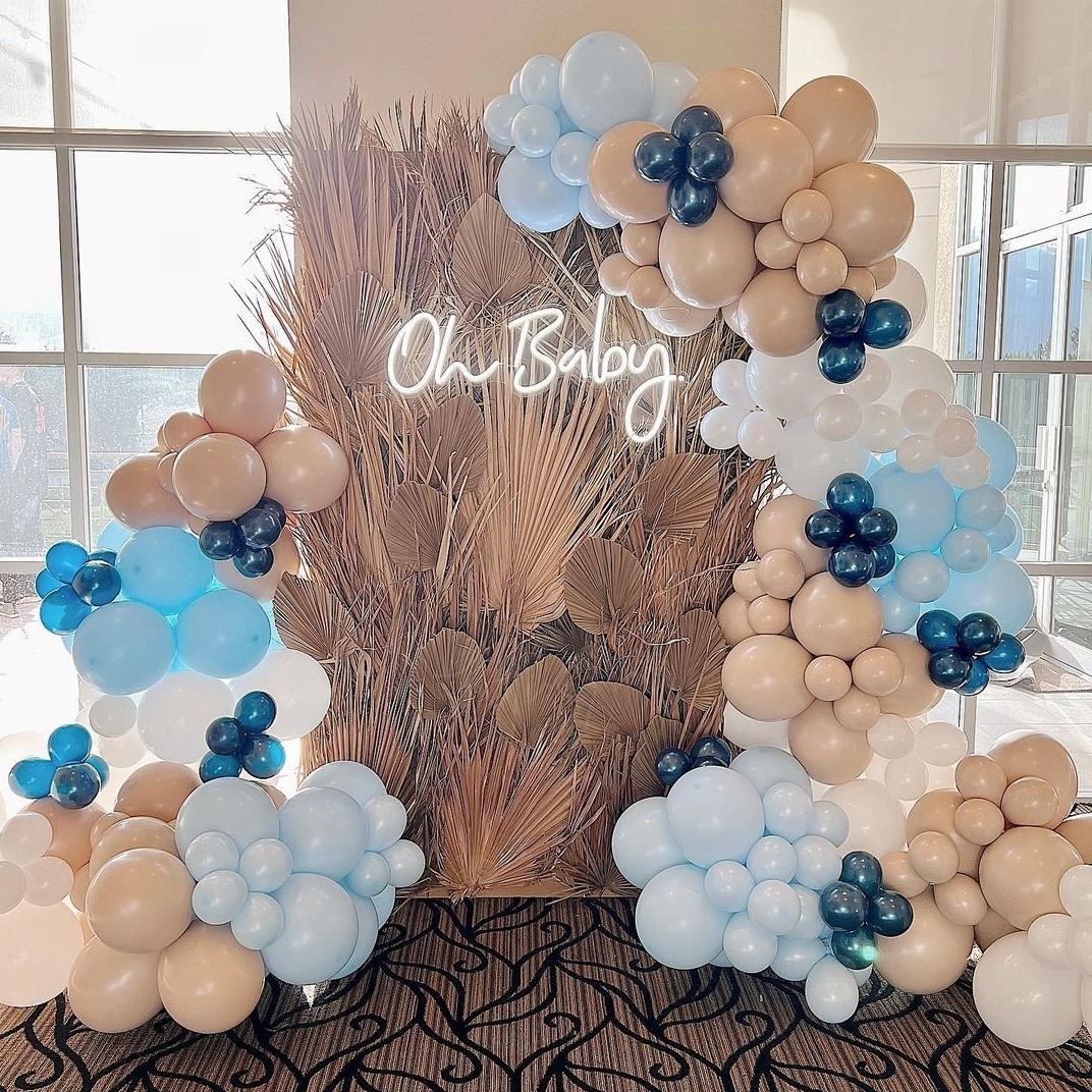 Dekoracja balonowa w odcieniach błękitu, beżu i bieli z ciemnoniebieskimi akcentami, tworząca łuk wokół ściany z suszonych traw i neonu 'Oh Baby', na tle okna.