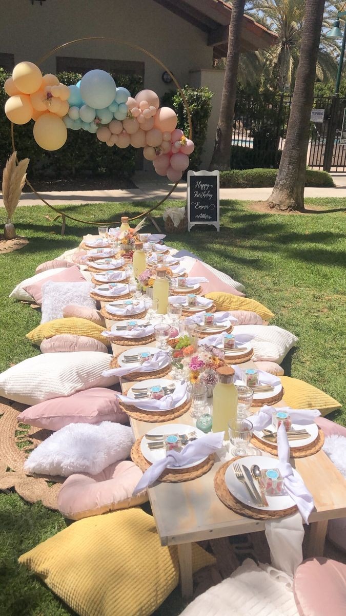 Uroczysty piknik urodzinowy na trawie z niskim stołem, poduszkami w pastelowych kolorach i dekoracją z balonów w kształcie łuku, tablica z napisem 'Happy Birthday Emma'.