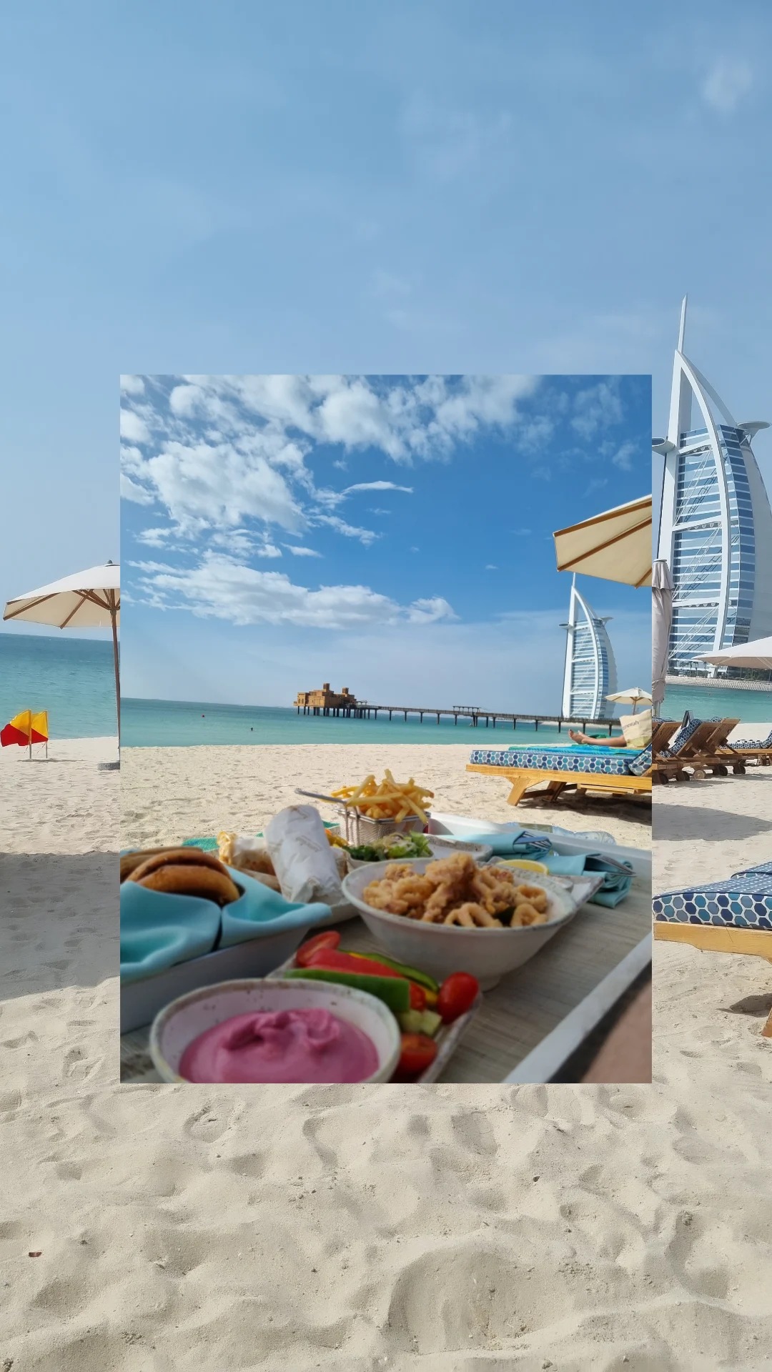 Luksusowy piknik na plaży w Dubaju z widokiem na Burj Al Arab: frytki, kalmary, warzywa i różowy dip na tle błękitnego nieba i turkusowej wody.