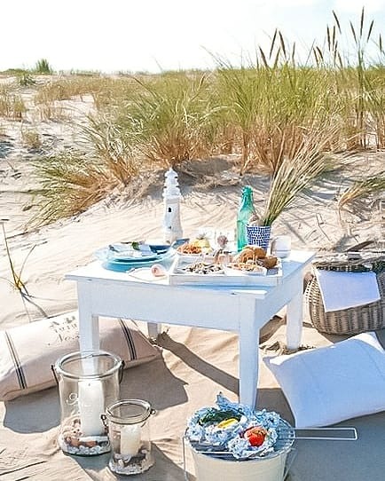 Piknik na plaży z białym stolikiem, dekoracjami marynistycznymi, koszem piknikowym, lampionami ze świecami i przenośnym grillem z warzywami w folii aluminiowej na piasku i trawie.