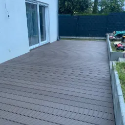 Nowoczesny taras z deski kompozytowej w odcieniach szarości, przylegający do białej ściany budynku z przesuwnymi drzwiami, widok na czarny płot i narzędzia w ogrodzie.