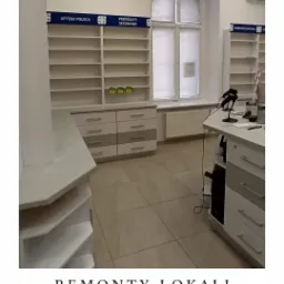 Wnętrze apteki po remoncie, widoczne nowe regały z oznaczeniami kategorii produktów, białe meble apteczne i okno z roletą.