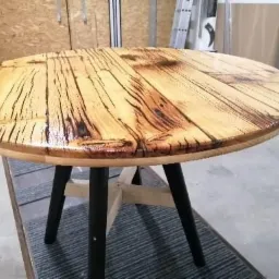 Okrągły blat stołu z jasnego drewna z ciemnymi żyłkami i czarnymi nogami, widoczne ślady lakierowania, w tle warsztat.