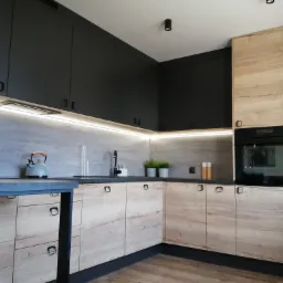 Nowoczesna kuchnia z drewnopodobnymi frontami szafek dolnych i czarnymi szafkami górnymi, podświetleniem LED blatu, czarnym blatem i elementami wykończeniowymi oraz sprzętem AGD.