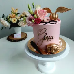 Różowy tort z imieniem 'Głosia', dekoracjami w kolorze złota: skrzydełka, aparat fotograficzny, czarne okulary, oraz kompozycją z różowych i białych kwiatów, umieszczony na białym stojaku, w tle...