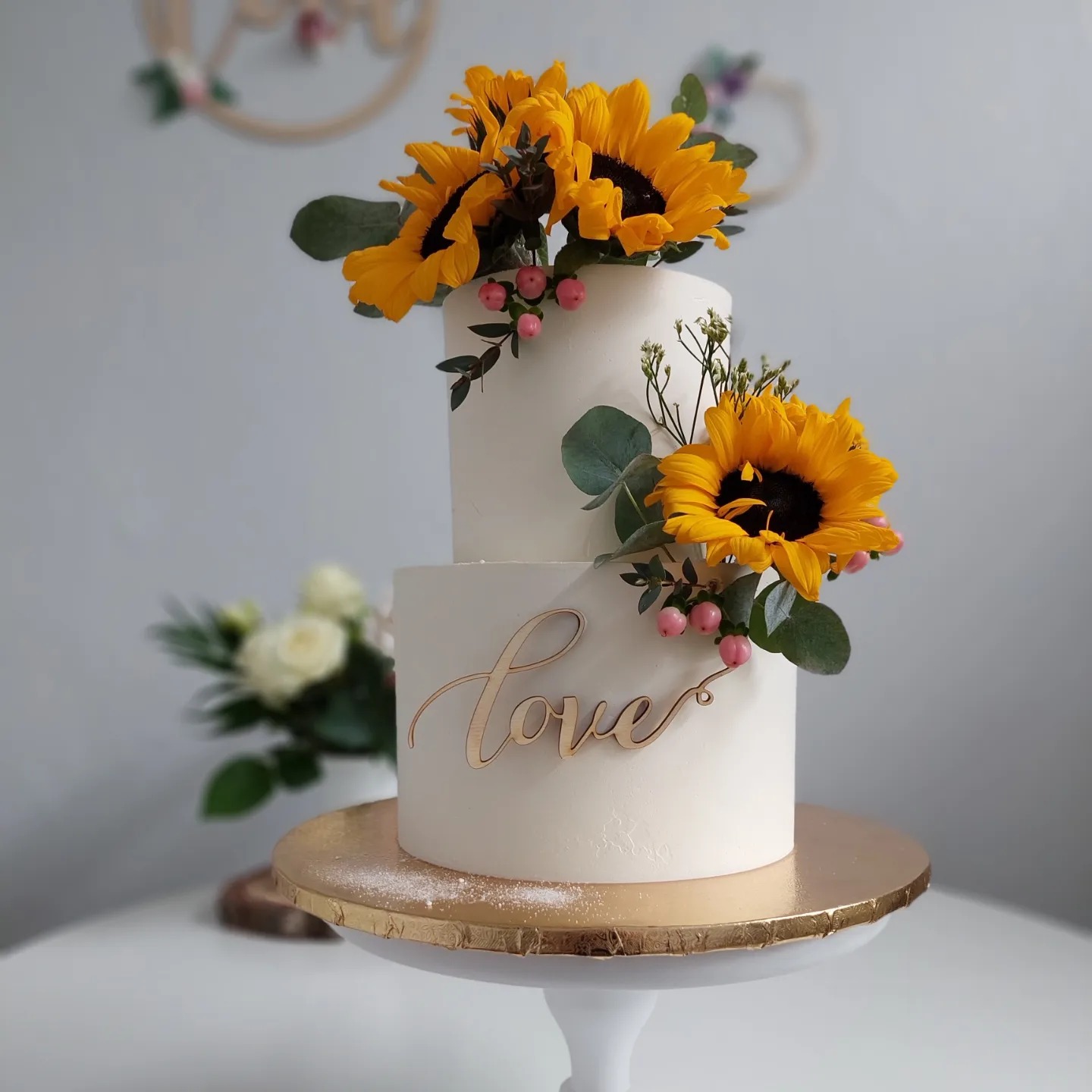 Dwupoziomowy tort weselny z białym lukrem, dekorowany świeżymi słonecznikami, eukaliptusem, drobnymi różowymi owocami i napisem 'love' z drewna, na złotym podeście.