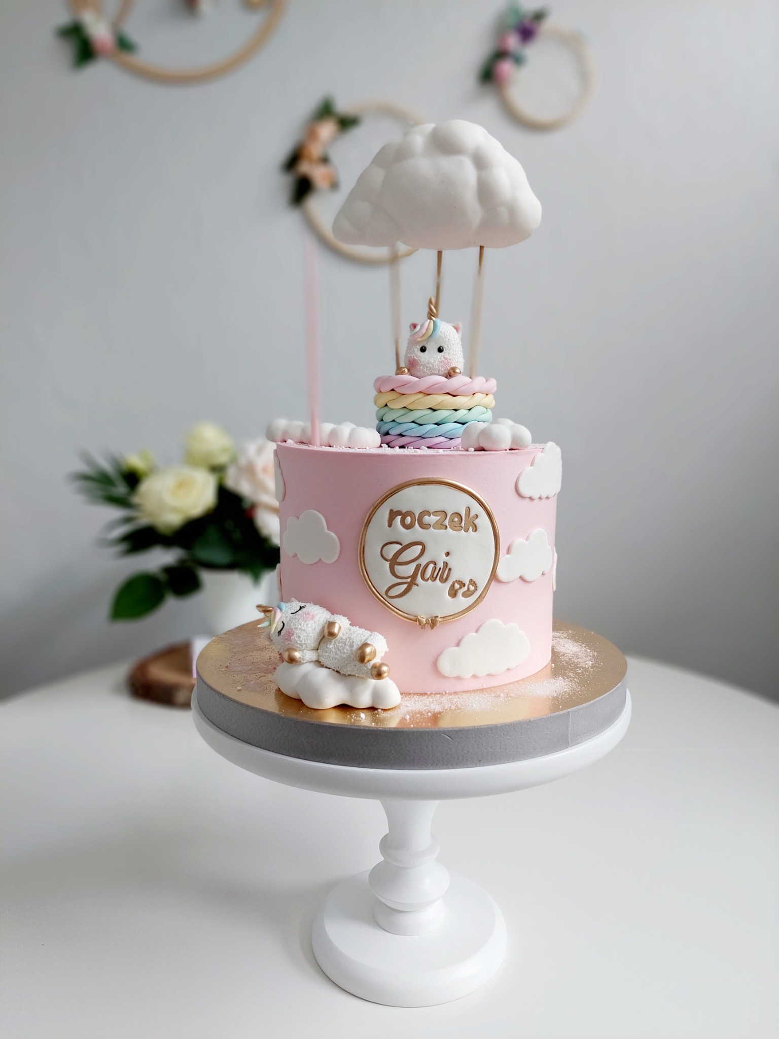 Różowy tort urodzinowy z napisem 'roczek Gai' otoczonym złotą ramką, ozdobiony chmurkami i figurkami jednorożców, umieszczony na białym stojaku na tle kwiatów i dekoracji ściennych.