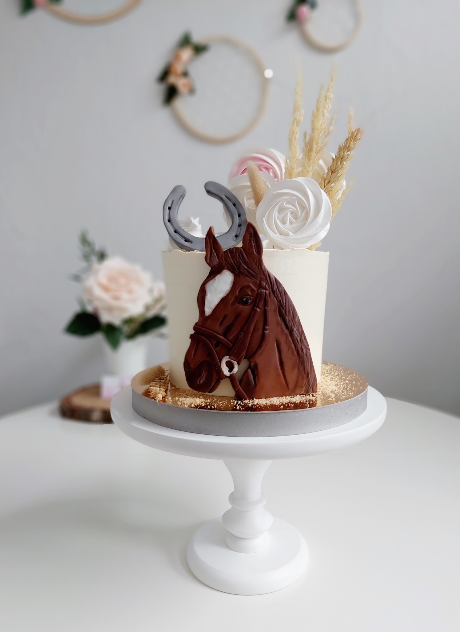 Tort z motywem konia i podkowy, dekorowany bezami i suszonymi trawami, umieszczony na białym stojaku.