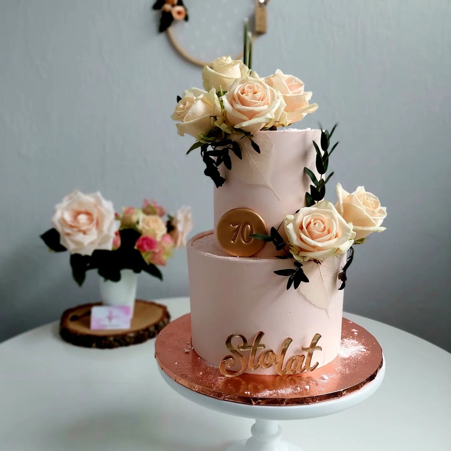 Dwupoziomowy tort w pudrowym różu, udekorowany kremowymi różami i zielonymi listkami, z topperem '70' i napisem 'Stolat' w złotym kolorze, umieszczony na białym stojaku na tle jasnoszarej ściany...