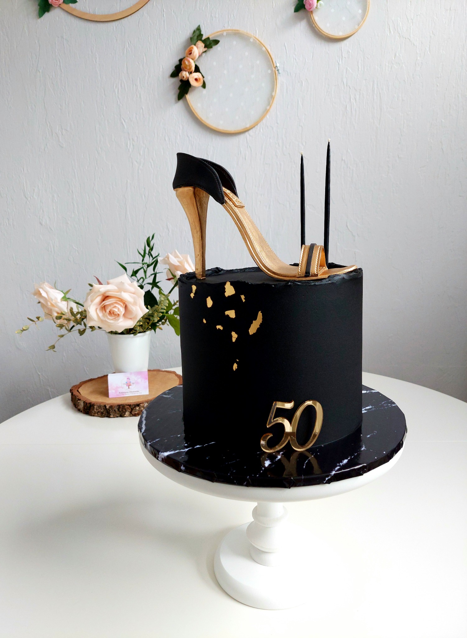 Czarny tort urodzinowy z dekoracją w kształcie złotego szpilki, numerem 50 i trzema czarnymi świeczkami, ustawiony na marmurowym paterze. W tle dekoracje ścienne i bukiet róż.
