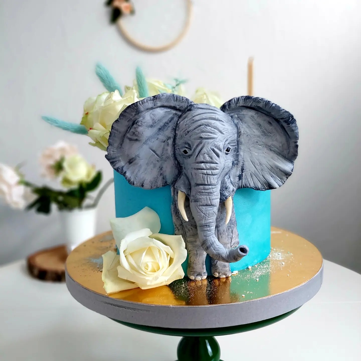 Turkusowy tort z szarą figurką słonia i białymi różami, umieszczony na złotym stojaku z szarym brzegiem i zieloną nóżką. W tle wazon z białymi kwiatami i dekoracyjny wianek.