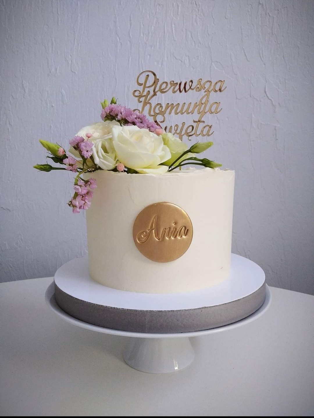 Tort komunijny z białym kremem, dekorowany świeżymi kwiatami i złotym topperem z napisem 'Pierwsza Komunia Święta' oraz złotym medalionem z imieniem 'Ania', umieszczony na białym stojaku.
