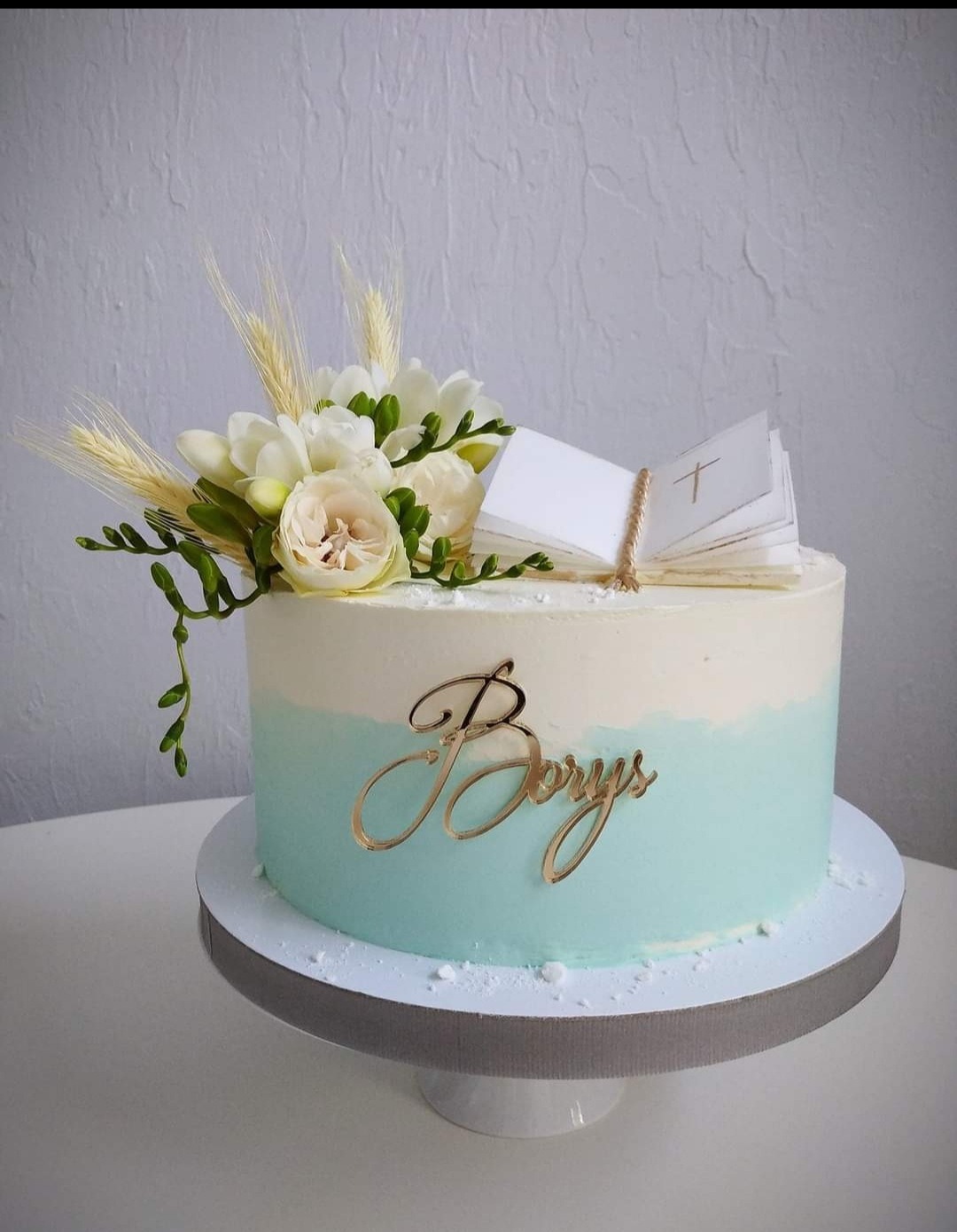 Tort ombre w kolorze błękitu i bieli, udekorowany kompozycją z białych kwiatów, źdźbeł zboża i miniaturowej książeczki z symbolem krzyża, z napisem 'Borys' wykonanym złotą czcionką.