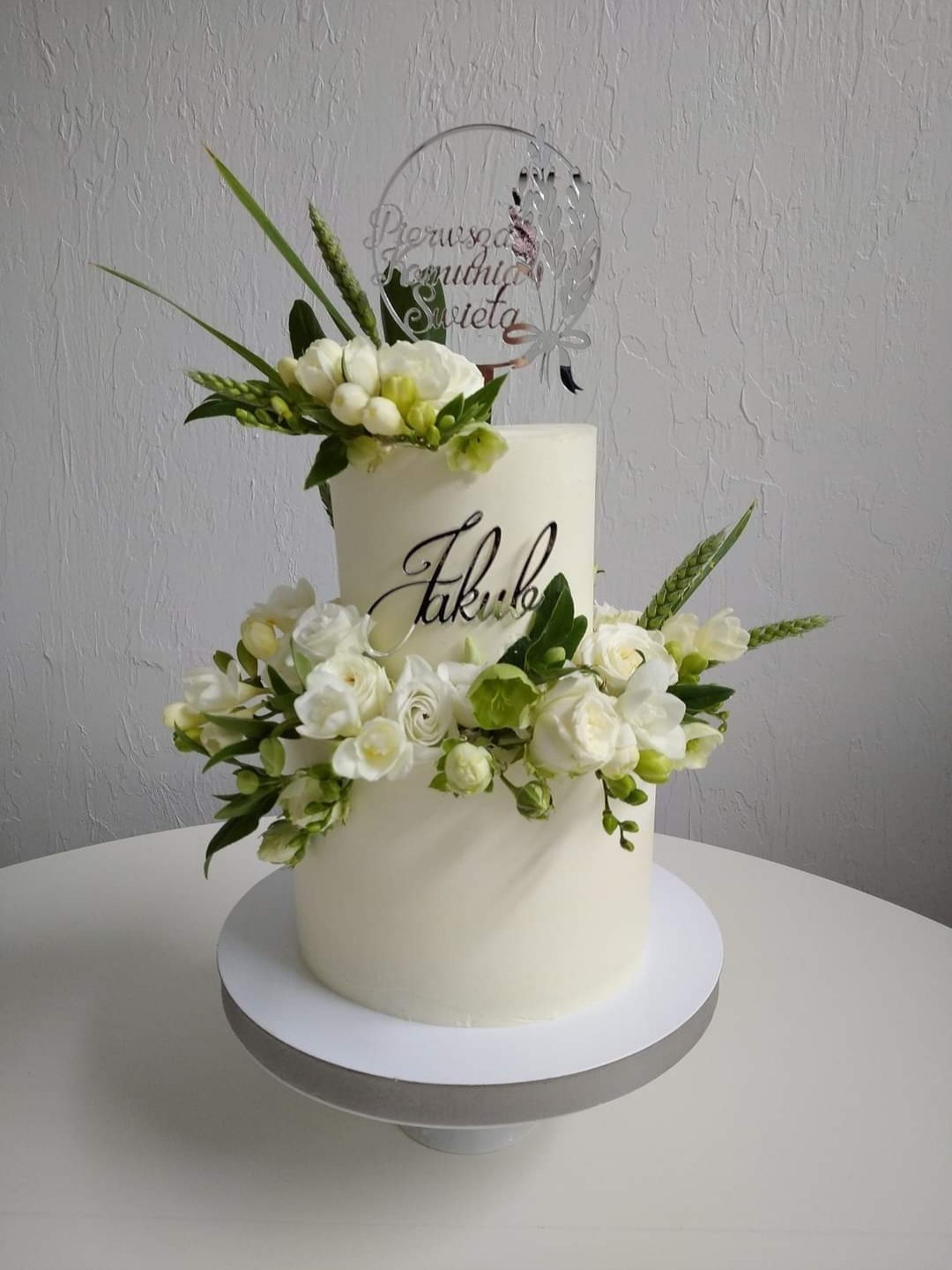 Dwupiętrowy tort komunijny z białym kremem, dekorowany świeżymi białymi kwiatami i topperem z napisem 'Pierwsza Komunia Święta' oraz imieniem 'Jakub' napisanym elegancką czcionką.