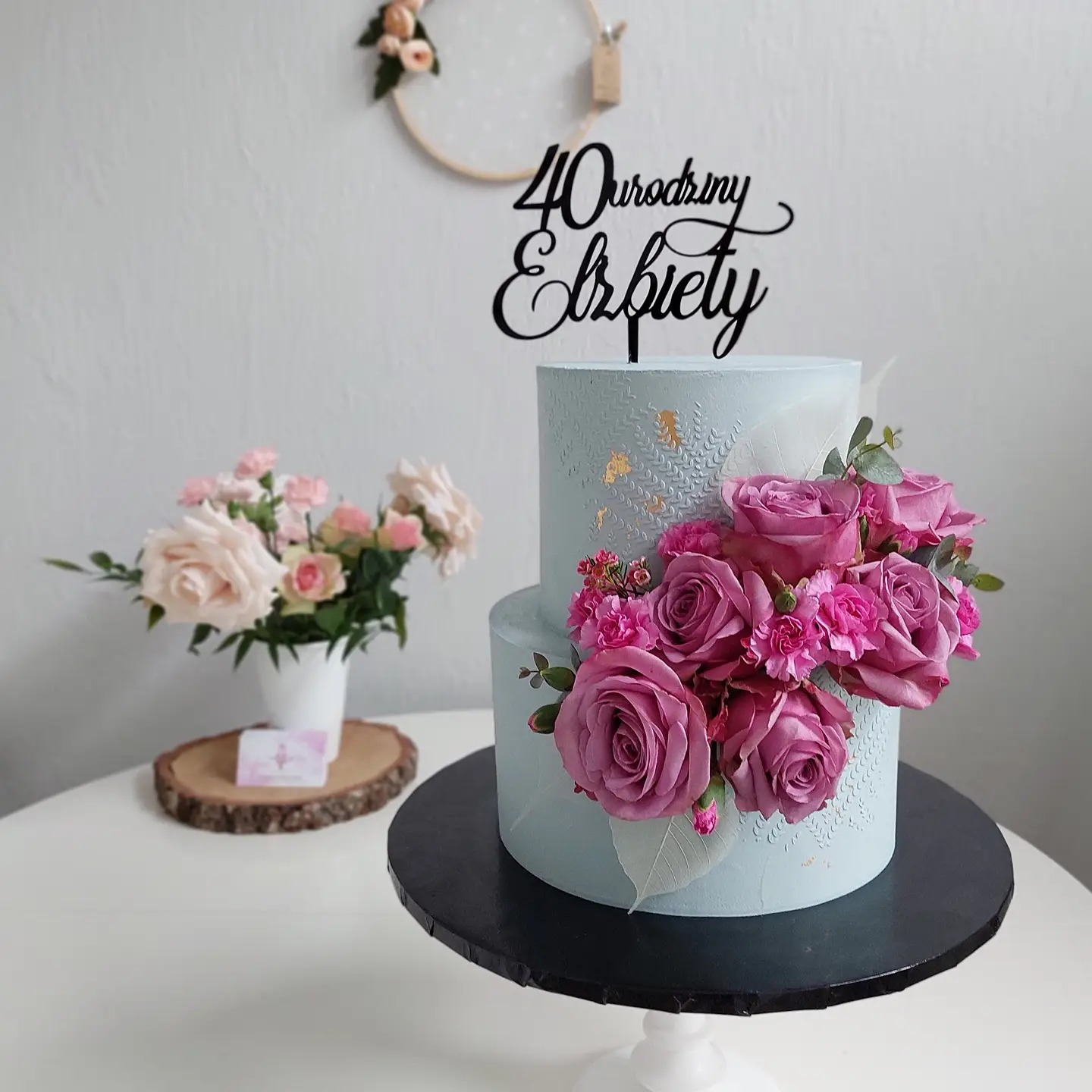 Dwupoziomowy tort w pastelowym błękicie, udekorowany różowymi różami i topperem z napisem '40 Urodziny Elżbiety', na białym stole, w tle bukiet różowych kwiatów i dekoracyjny wieniec.