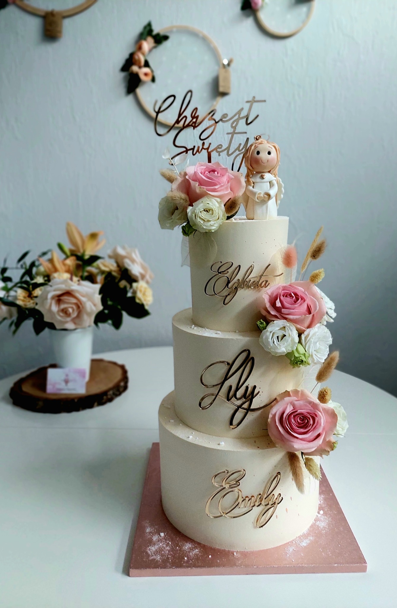 Trzypiętrowy tort w kremowym kolorze, udekorowany różowymi i białymi różami, imionami z akrylu w kolorze złota (Elżbieta, Lily, Emily) oraz topperem z napisem 'Chrzest Święty' i figurką aniołka,...