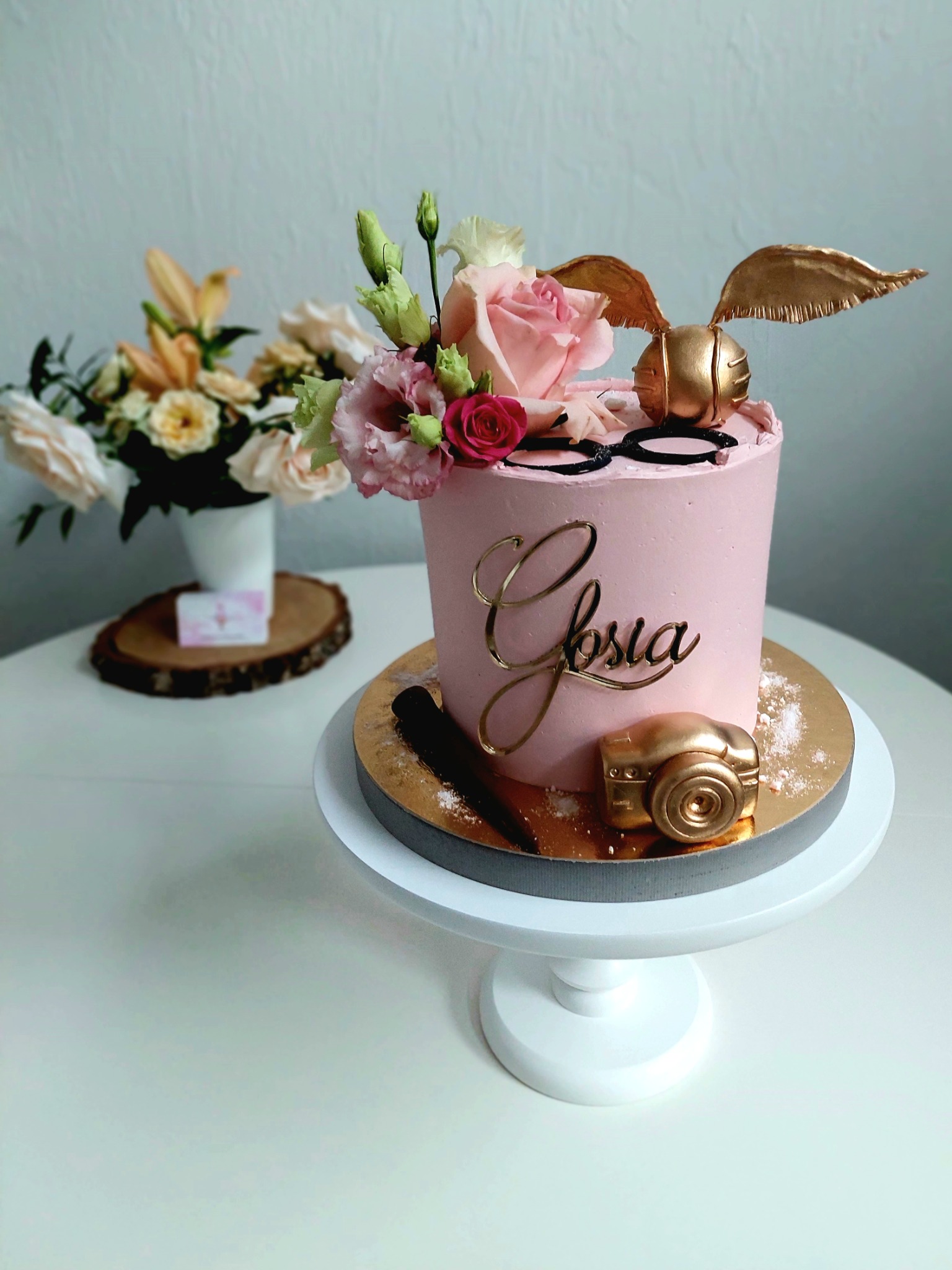 Różowy tort z imieniem 'Głosia', dekoracjami w kolorze złota: skrzydełka, aparat fotograficzny, czarne okulary, oraz kompozycją z różowych i białych kwiatów, umieszczony na białym stojaku, w tle...