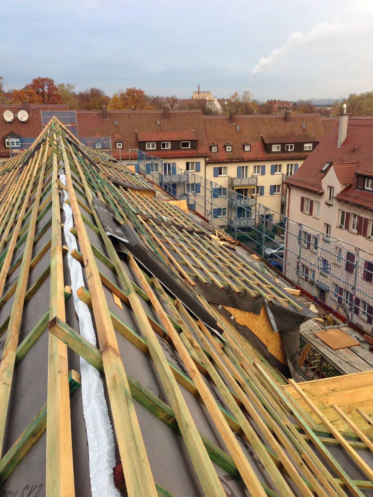 Konstrukcja dachu w trakcie budowy z widocznymi łatami, membraną i elementami izolacji, na tle zabudowy miejskiej z czerwonymi dachami i kominami.