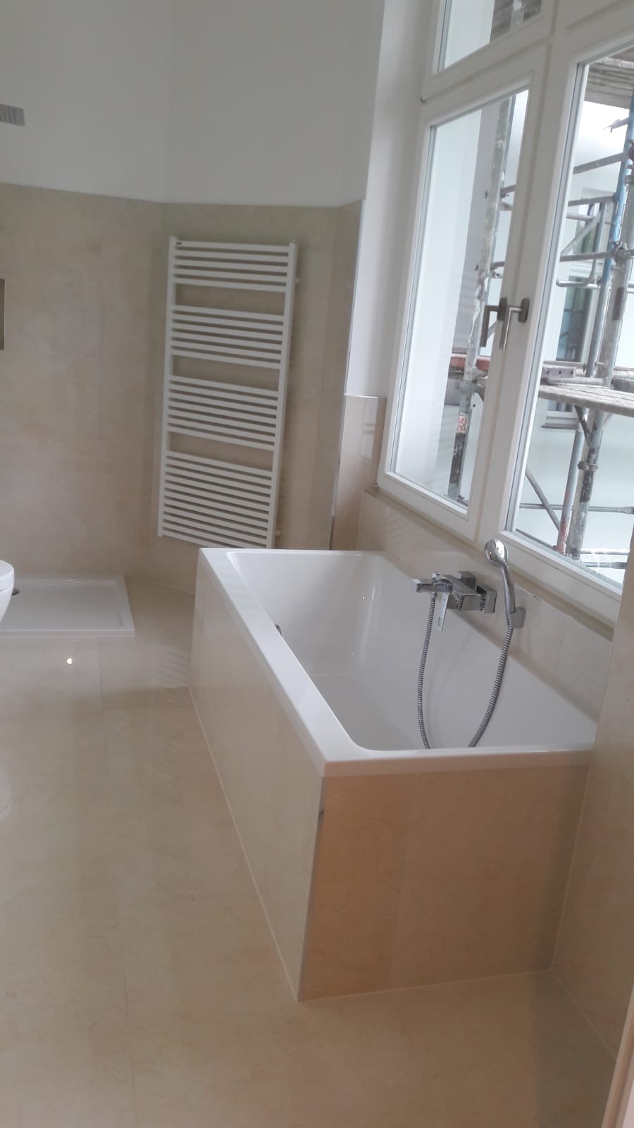 Wykończona łazienka z beżowymi płytkami, białą wanną i grzejnikiem drabinkowym; za oknem widoczne rusztowania.