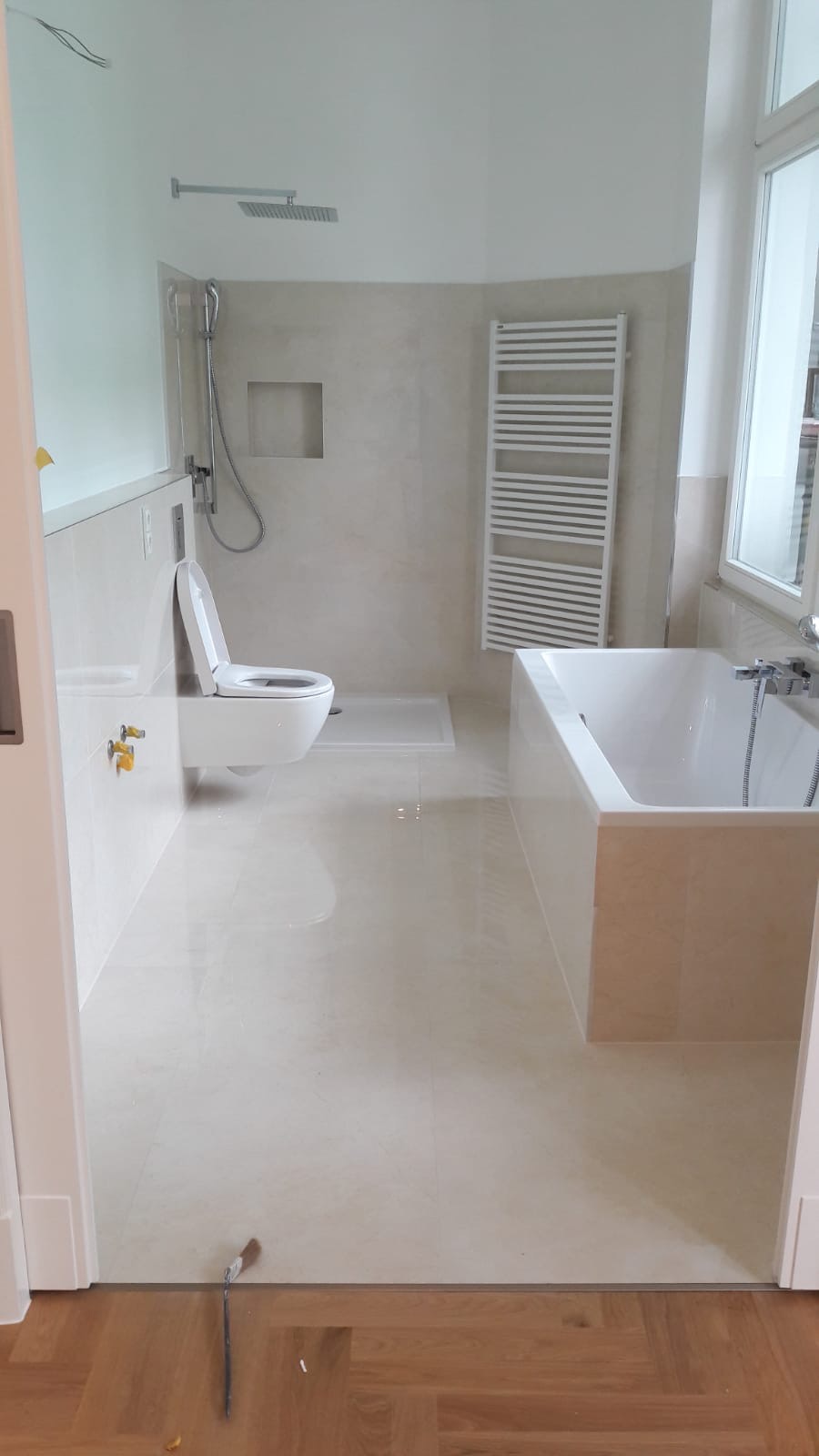 Jasna łazienka po remoncie z beżowymi płytkami, wiszącą toaletą, grzejnikiem drabinkowym i kabiną prysznicową typu walk-in, widok zza progu z parkietem i pędzlem malarskim.