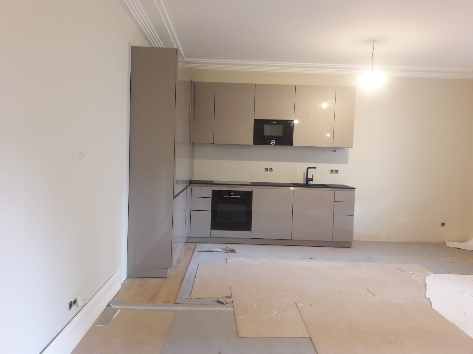 Nowoczesna kuchnia z beżowymi szafkami na wysoki połysk, czarnym blatem i sprzętem AGD, widoczna na etapie prac wykończeniowych, z zabezpieczoną podłogą.