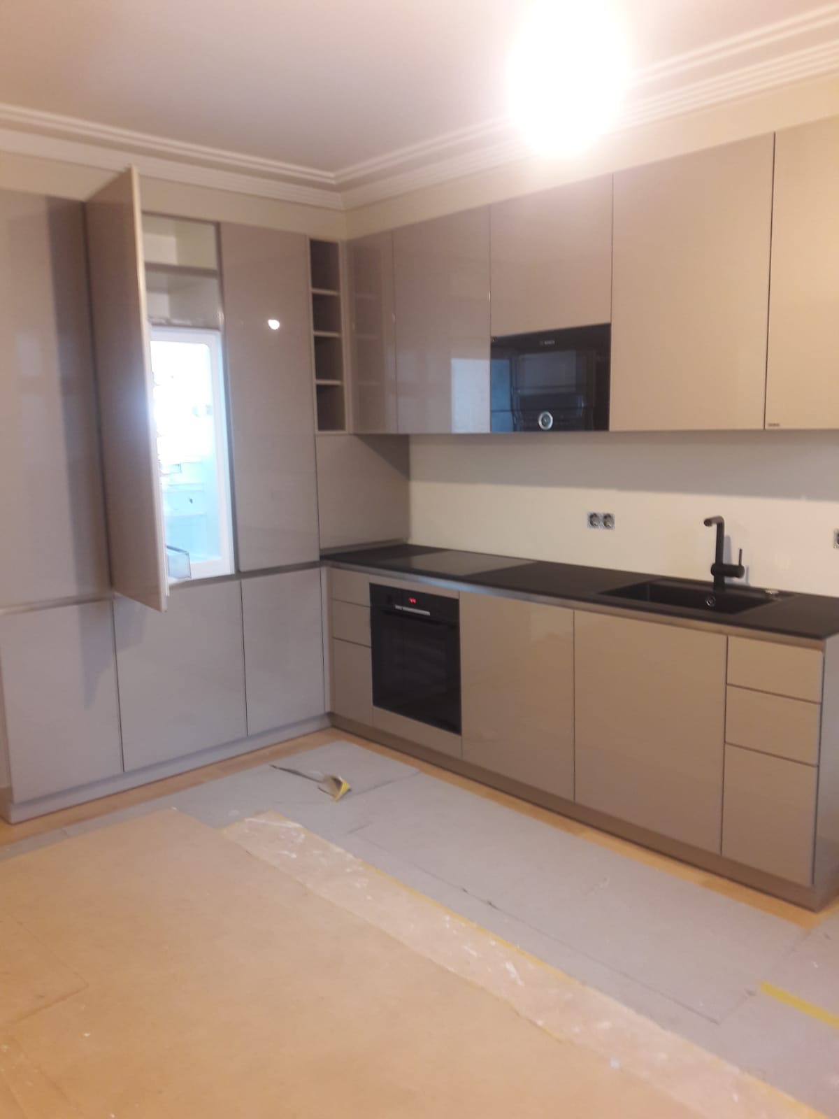 Nowoczesna kuchnia z beżowymi, lakierowanymi frontami szafek, czarnym blatem i sprzętem AGD w zabudowie. Na podłodze widoczne płyty kartonowo-gipsowe, prawdopodobnie przygotowane do układania podłogi.