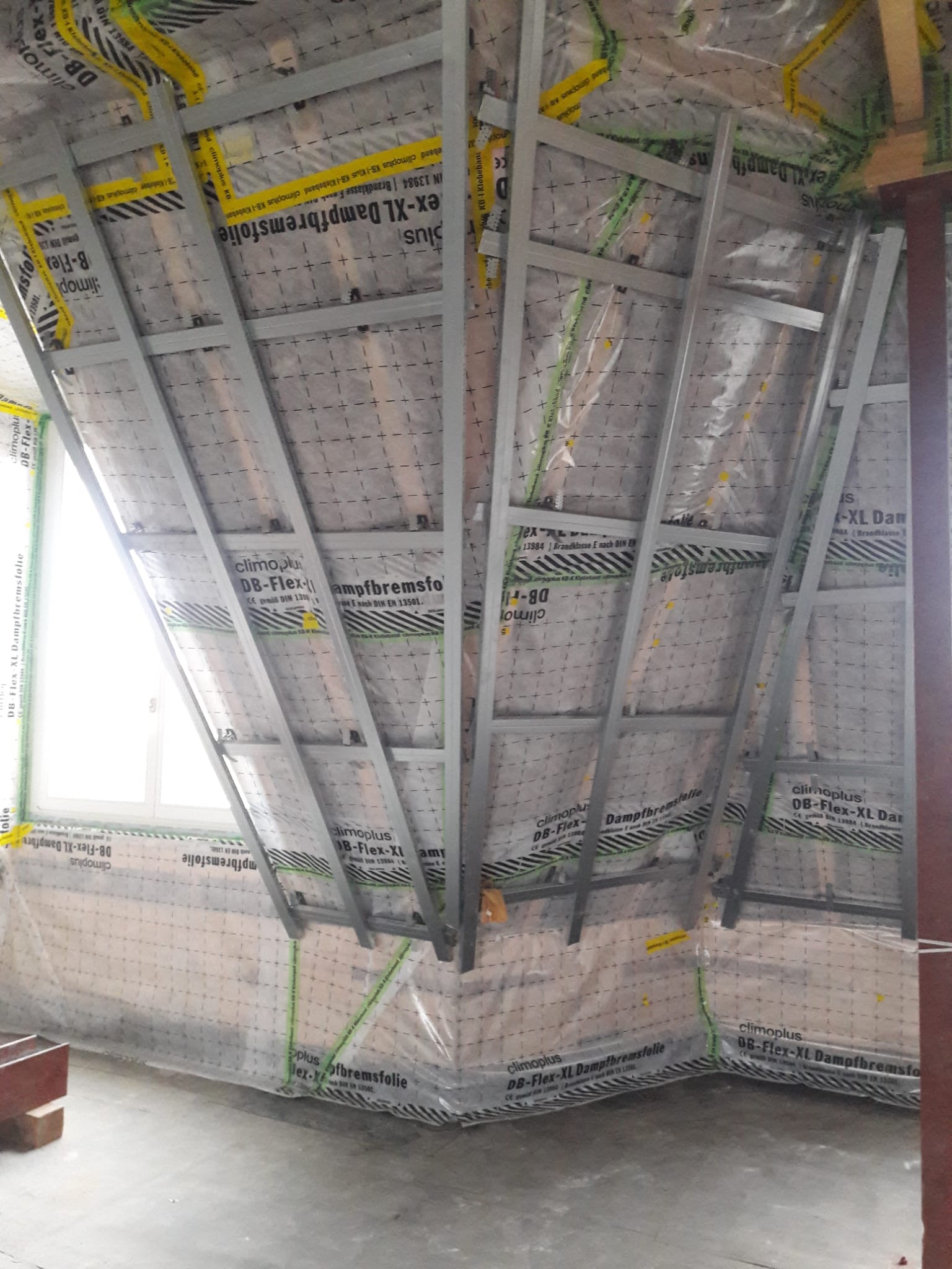 Montaż stelaża pod płytę gipsową na poddaszu, widoczna folia paroizolacyjna z nadrukiem, okno w tle.