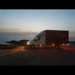 Biały samoch&oacute;d dostawczy z czarną plandeką zaparkowany na klifie z widokiem na morze o zmierzchu. Włączone światła przednie i tylne.