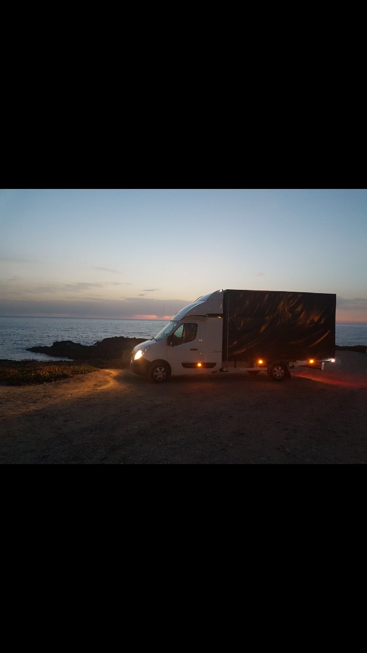 Biały samochód dostawczy z czarną plandeką zaparkowany na klifie z widokiem na morze o zmierzchu. Włączone światła przednie i tylne.