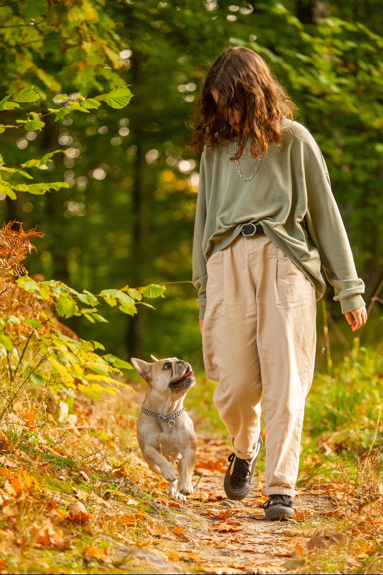 Młoda kobieta z kręconymi włosami i srebrnym łańcuszkiem na szyi spaceruje w lesie z buldogiem francuskim na smyczy. Pies biegnie pośród opadłych liści, spoglądając w górę na właścicielkę.