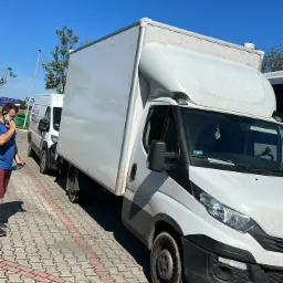 Biały samochód dostawczy typu Iveco z zabudową kontenerową na parkingu z kostki, częściowo zasłonięty przez osobę rozmawiającą przez telefon. Błękitne niebo w tle.