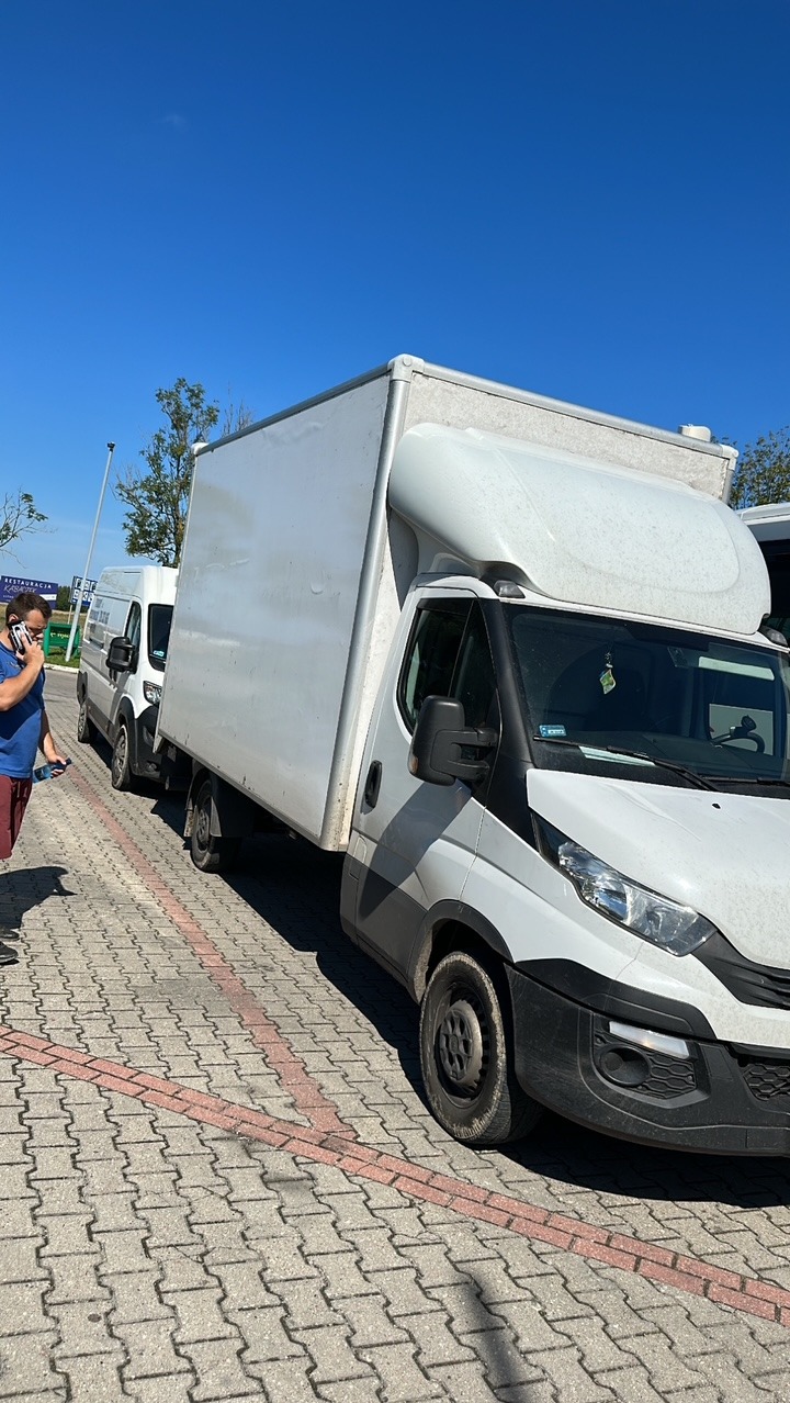 Biały samochód dostawczy typu Iveco z zabudową kontenerową na parkingu z kostki, częściowo zasłonięty przez osobę rozmawiającą przez telefon. Błękitne niebo w tle.