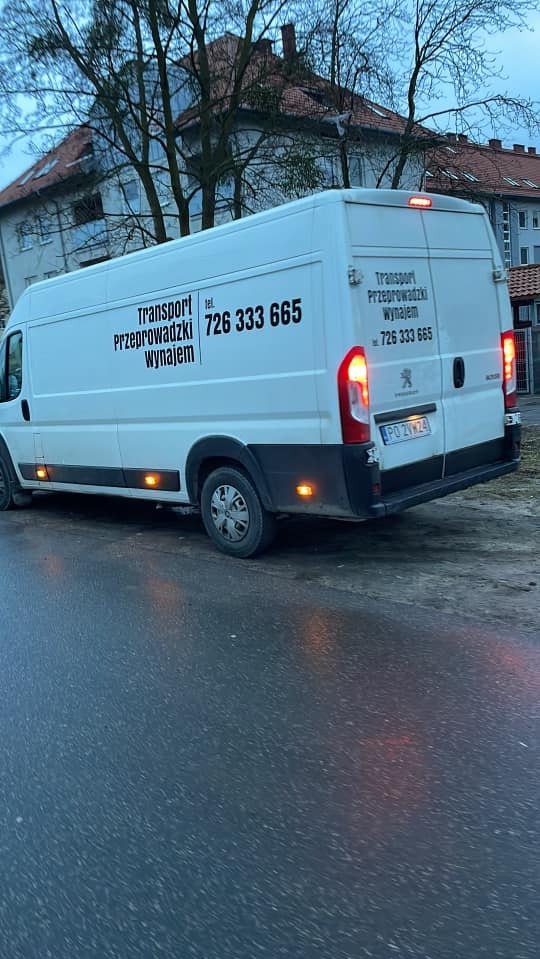 Biały bus transportowy z czarnymi napisami 'Transport Przeprowadzki Wynajem' i numerem telefonu 726 333 665, zaparkowany na mokrej ulicy w tle budynki.