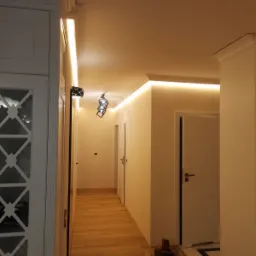 Korytarz z drewnianą podłogą i białymi ścianami, oświetlony listwami LED pod sufitem i wiszącą lampą o geometrycznym kształcie. Widoczne narzędzia na podłodze sugerują trwające prace wykończeniowe.