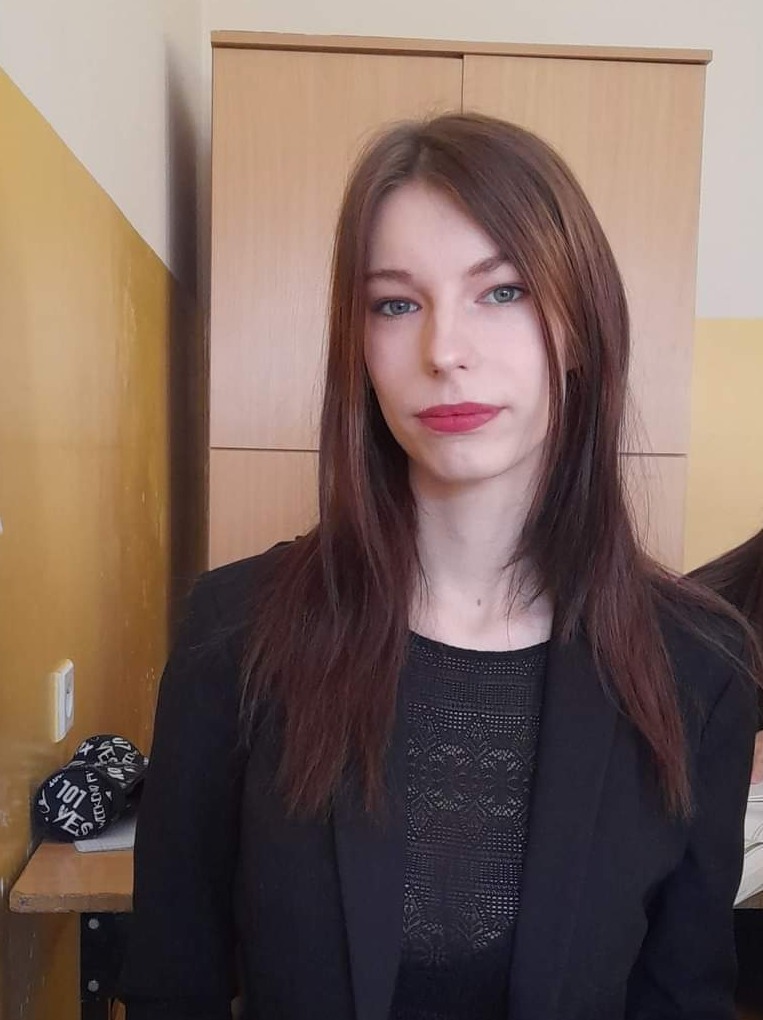 Portret młodej kobiety z długimi brązowymi włosami, ubranej w czarny żakiet i koronkową bluzkę, na tle ściany z szafką.