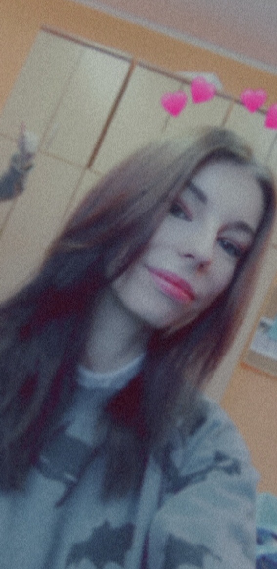 Portret kobiety z długimi brązowymi włosami, uśmiechającej się do kamery, z filtrem różowych serduszek nad głową, w szarej bluzie moro na tle beżowej ściany.