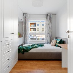 Joanna Lar - Plena Invest - Przytulna sypialnia w Łodzi z dużym, białym wbudowanym szafą i łóżkiem z zielonym pledem. Widok z okna na budynek mieszkalny. Aranżacja wnętrza.