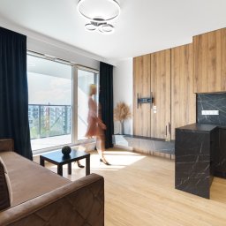 Joanna Lar - Plena Invest - Nowoczesne wnętrze: sofa, stół, zabudowa drewniana z TV, marmurowy blat kuchenny. Rozmyta sylwetka kobiety przechodzącej obok balkonu. Stylowe oświetlenie sufitowe.