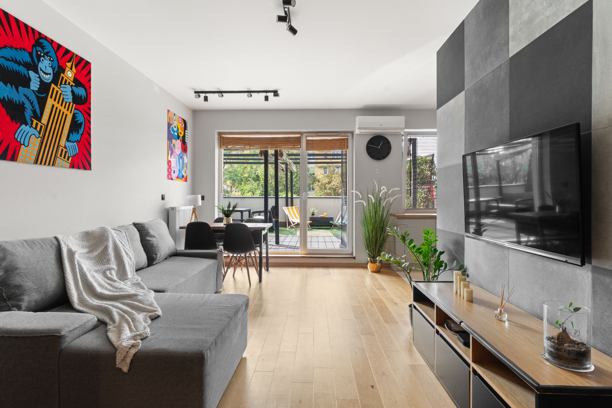 Jasne wnętrze mieszkania z szarą sofą, drewnianą podłogą i wyjściem na balkon. Na ścianie abstrakcyjne obrazy i telewizor na tle szarej ściany.