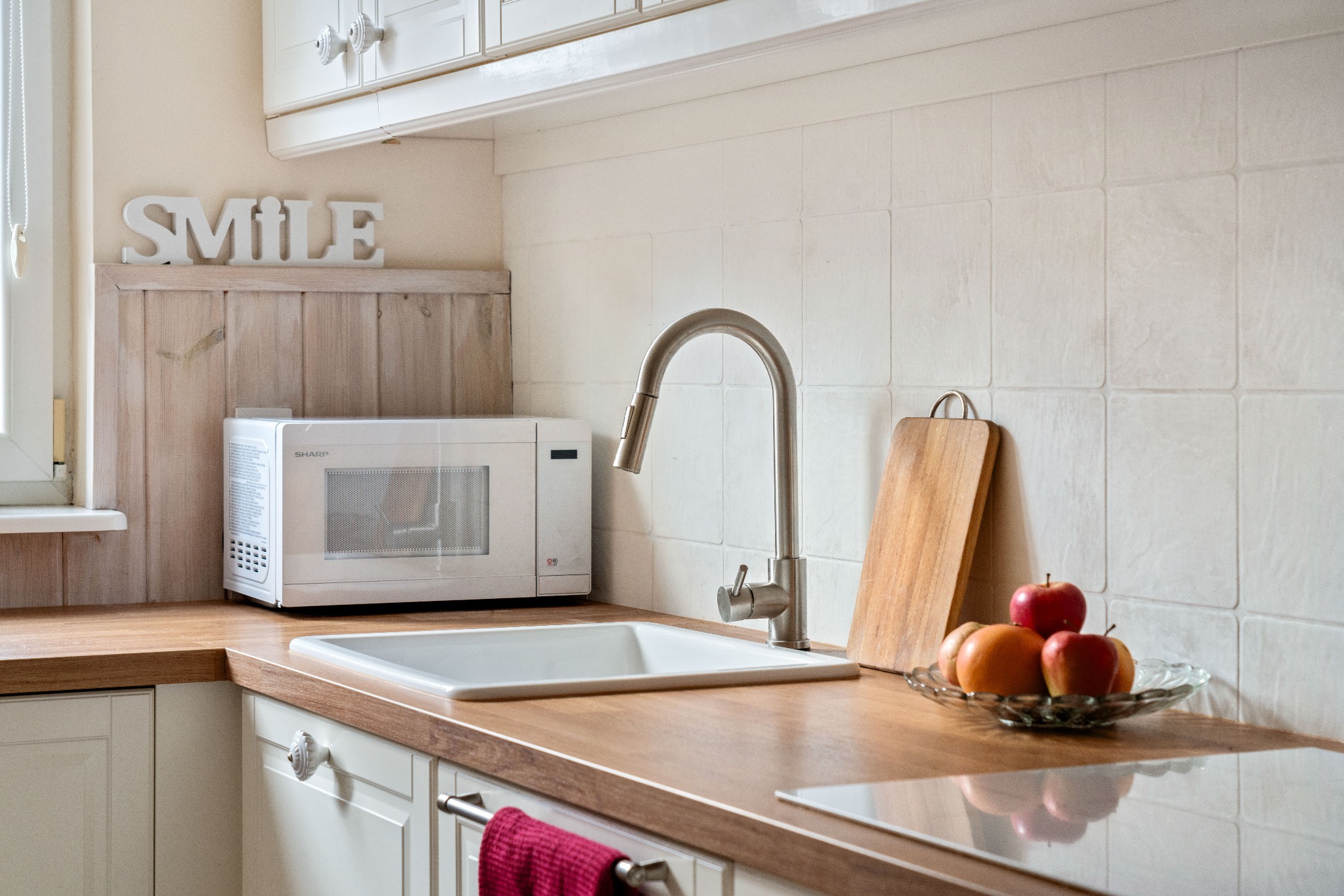 Jasna kuchnia z drewnianym blatem, owocami i napisem 'SMILE'. Ujęcie prezentuje aranżację wnętrza z bliska, z widoczną armaturą i wyposażeniem.