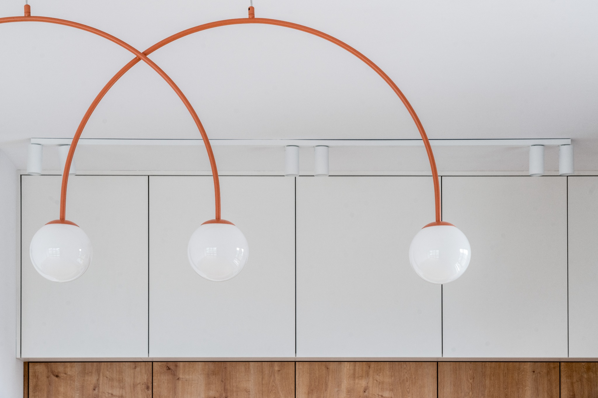 Nowoczesna kuchnia: białe szafki z drewnianym akcentem i designerskie lampy wiszące z pomarańczowymi łukami, oświetlające wnętrze.