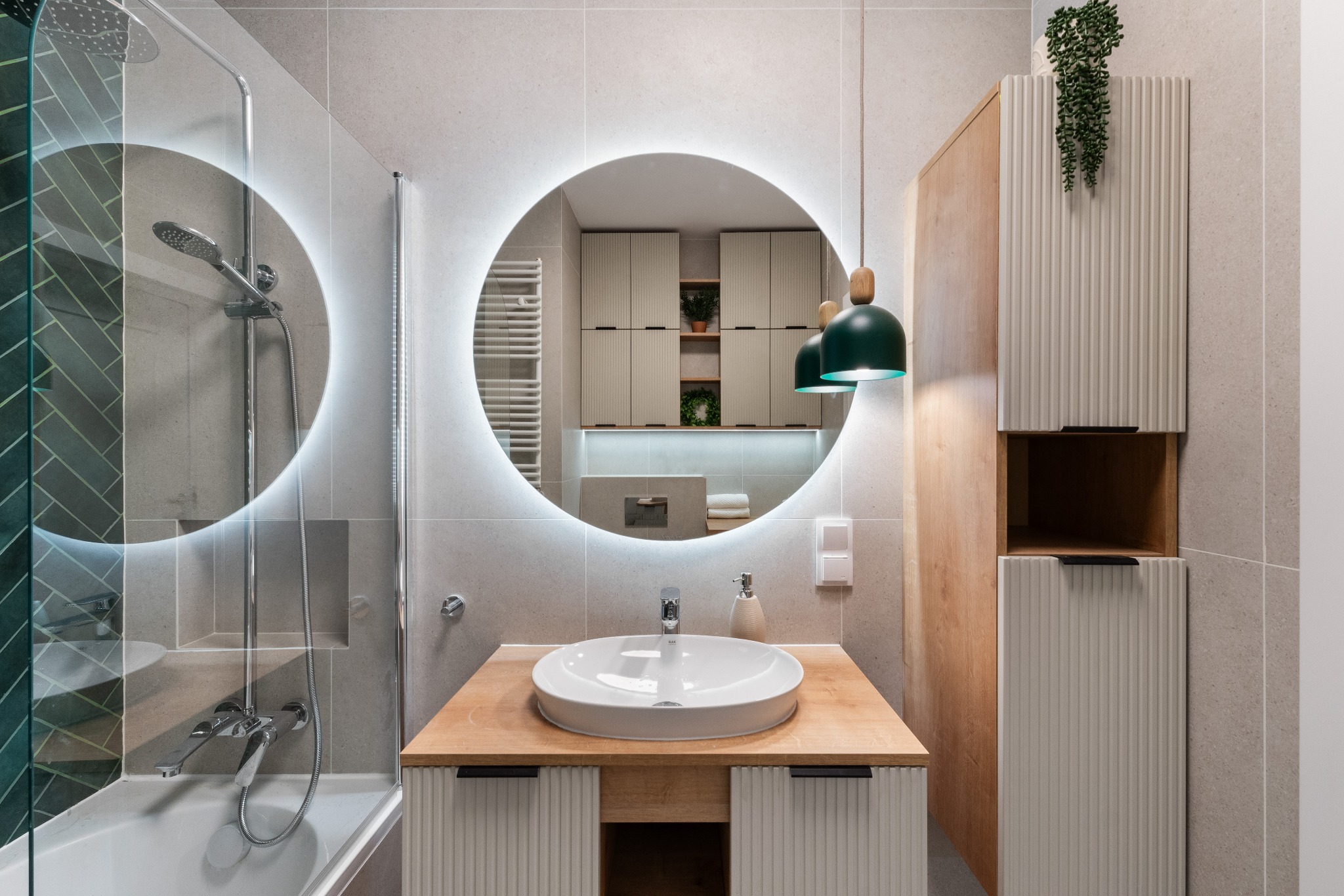 Nowoczesna łazienka z okrągłym lustrem podświetlanym LED, drewnianym blatem umywalki i kabiną prysznicową z zieloną mozaiką w jodełkę. Roślina zwisa z szafki.