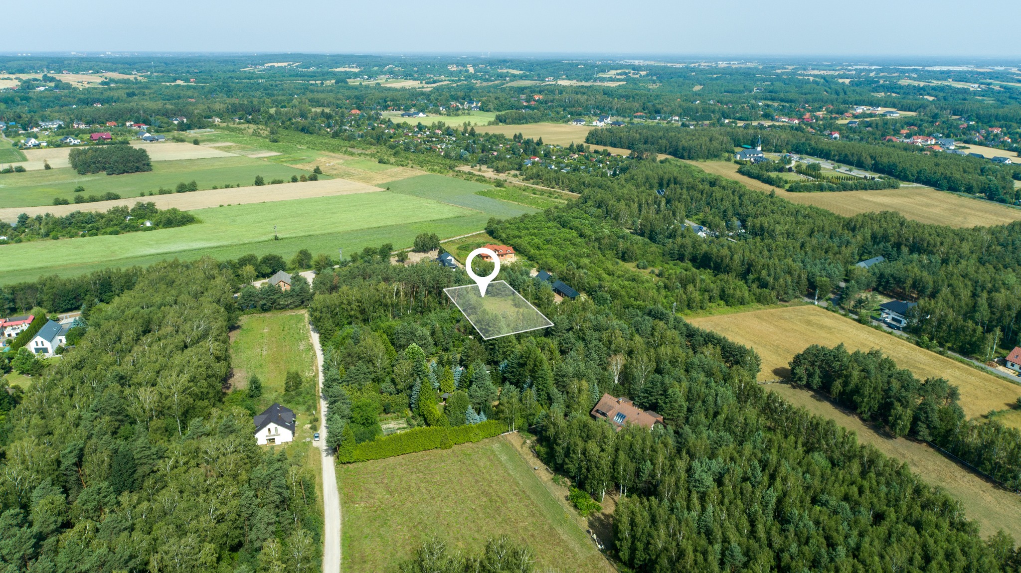 Zdjęcie z lotu ptaka przedstawia działkę budowlaną otoczoną lasem, z zaznaczoną granicą i pinezką lokalizacyjną. W tle widoczne domy i pola uprawne.
