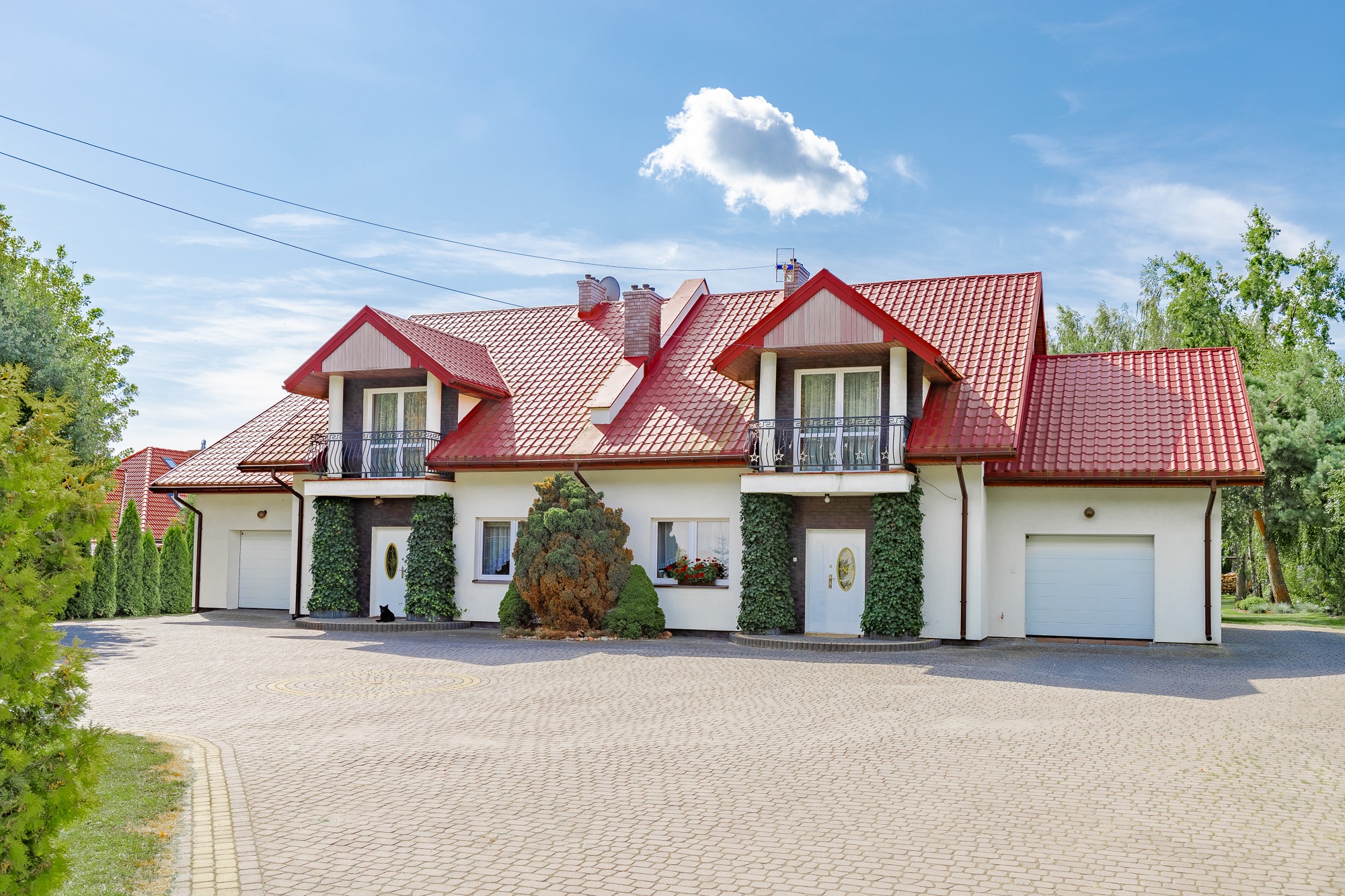 Biały dom dwurodzinny z czerwonym dachem i balkonami, porośnięty bluszczem, z brukowanym podjazdem i zadbanym ogrodem w słoneczny dzień.