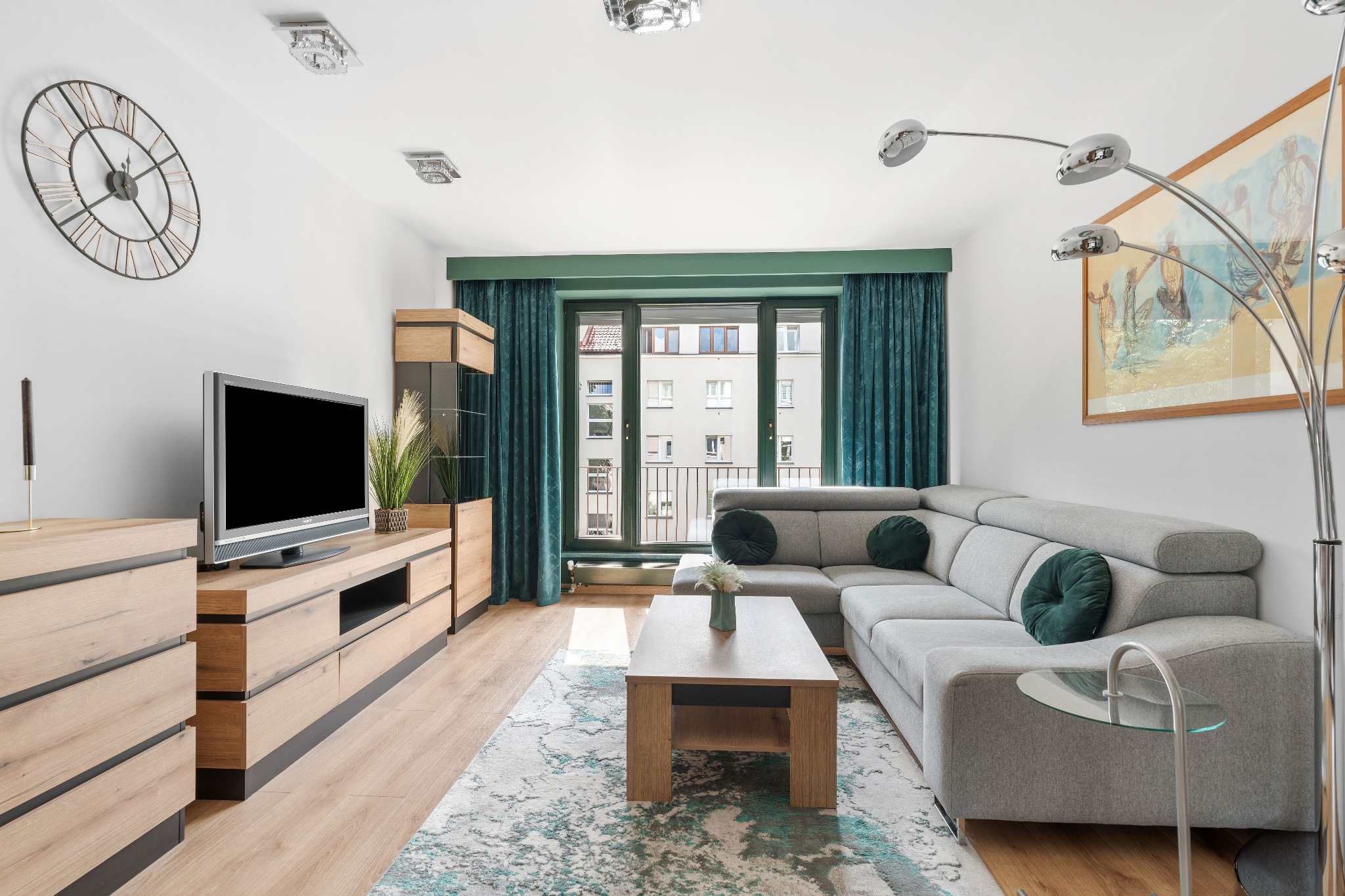 Nowoczesny salon z szarą sofą narożną, drewnianą komodą pod telewizor i turkusowymi zasłonami. Duży zegar na ścianie. Jasne, przestronne wnętrze z balkonem.
