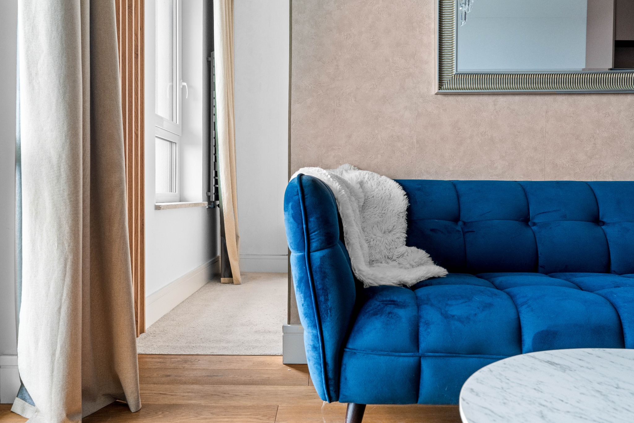 Nowoczesny salon: błękitna sofa z poduszką, marmurowy stolik, drewniana podłoga i okno z zasłonami tworzą przytulne, stylowe wnętrze.